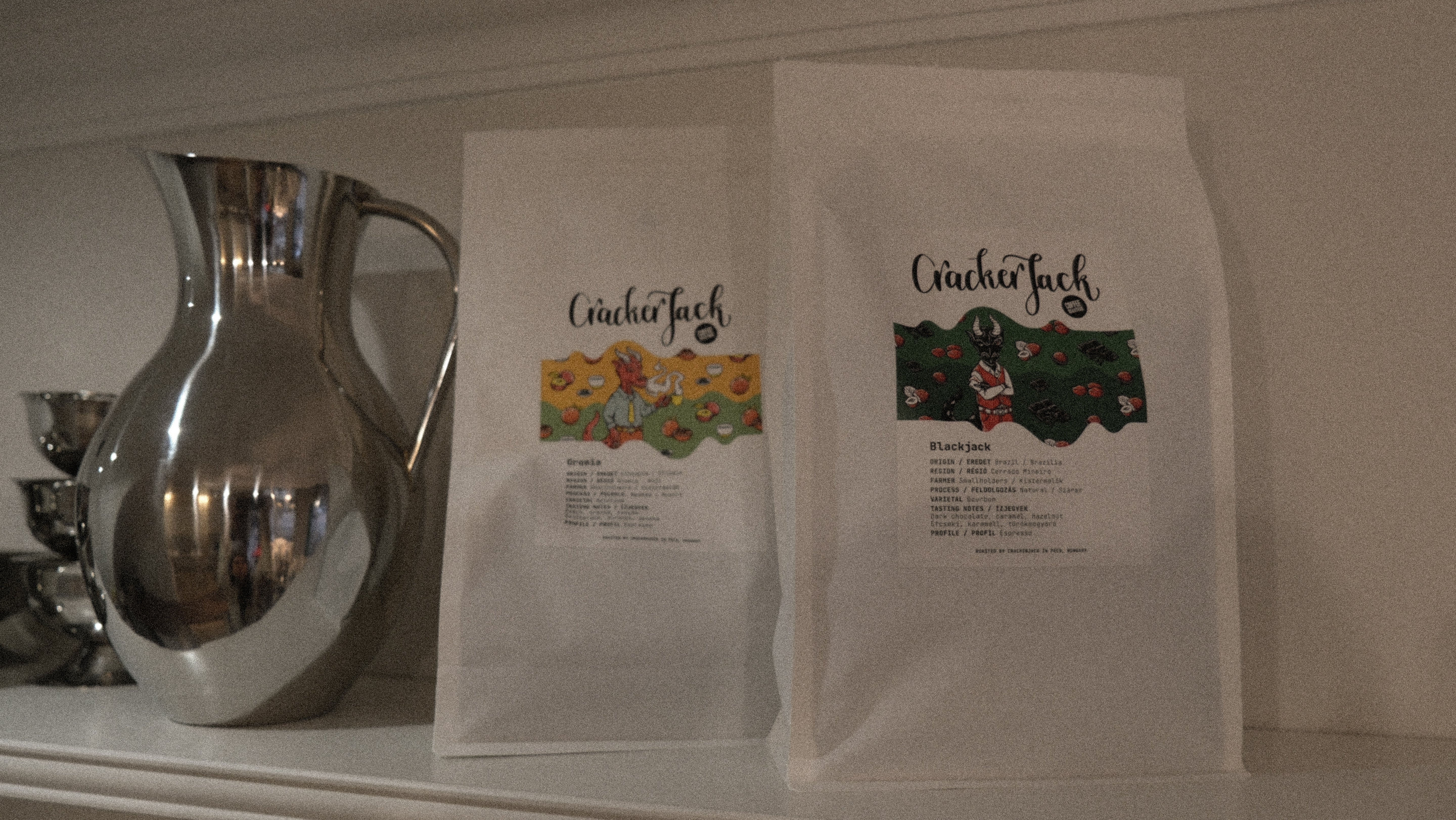 Two bags of Cracker Jack coffee on a white shelfe with a silver pitcher in the background.