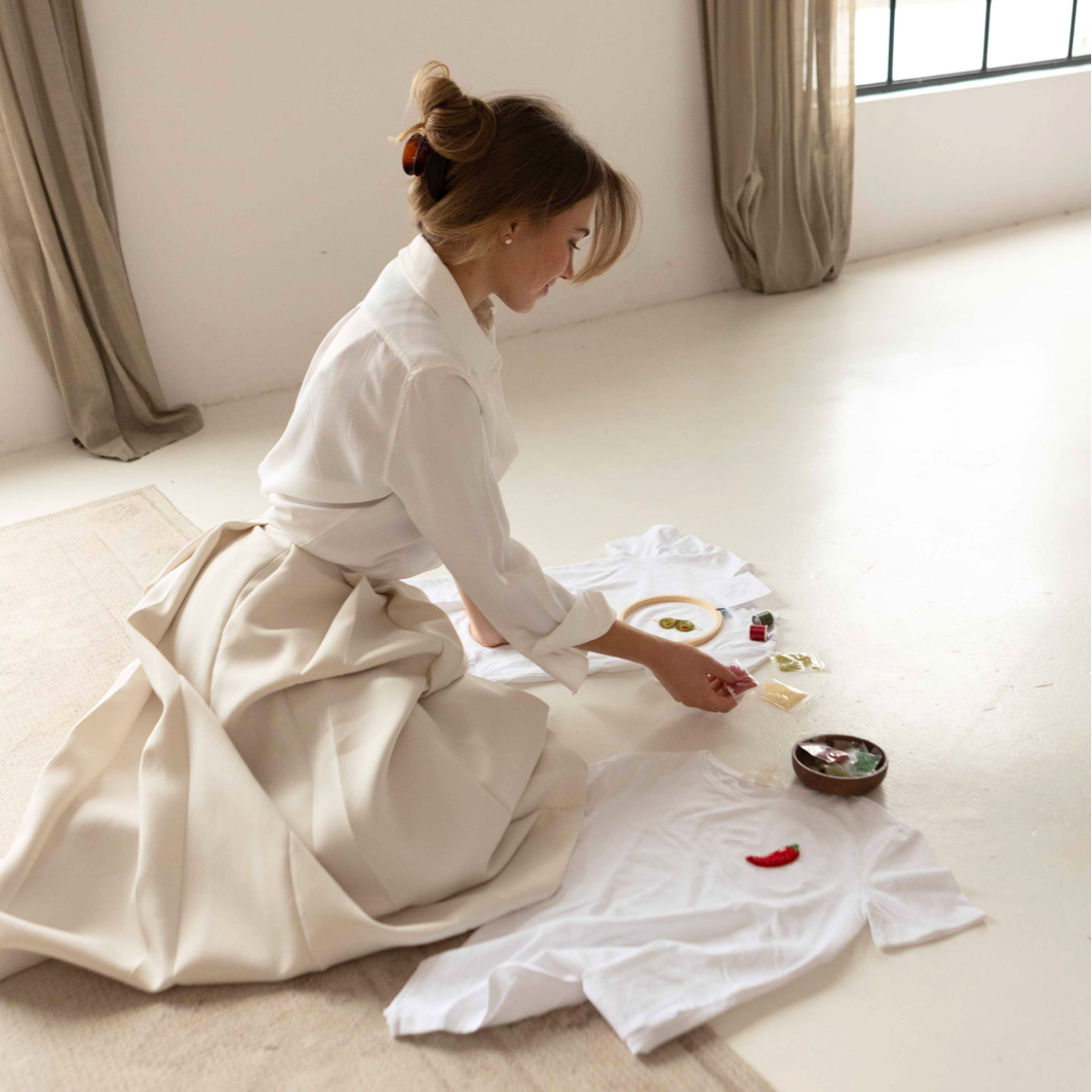 Woman sitting on the floor in elegant cream outfit, working on DIY beaded embroidery t-shirts with colorful seed beads and hoops.