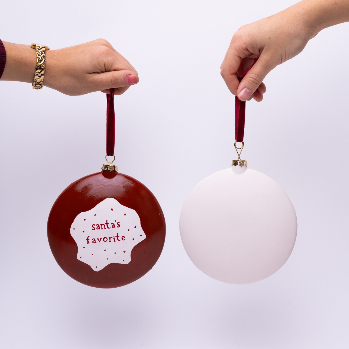 Two Christmas ornaments, one red with 'Santa's favorite' text and one white, held by hands against a light background.