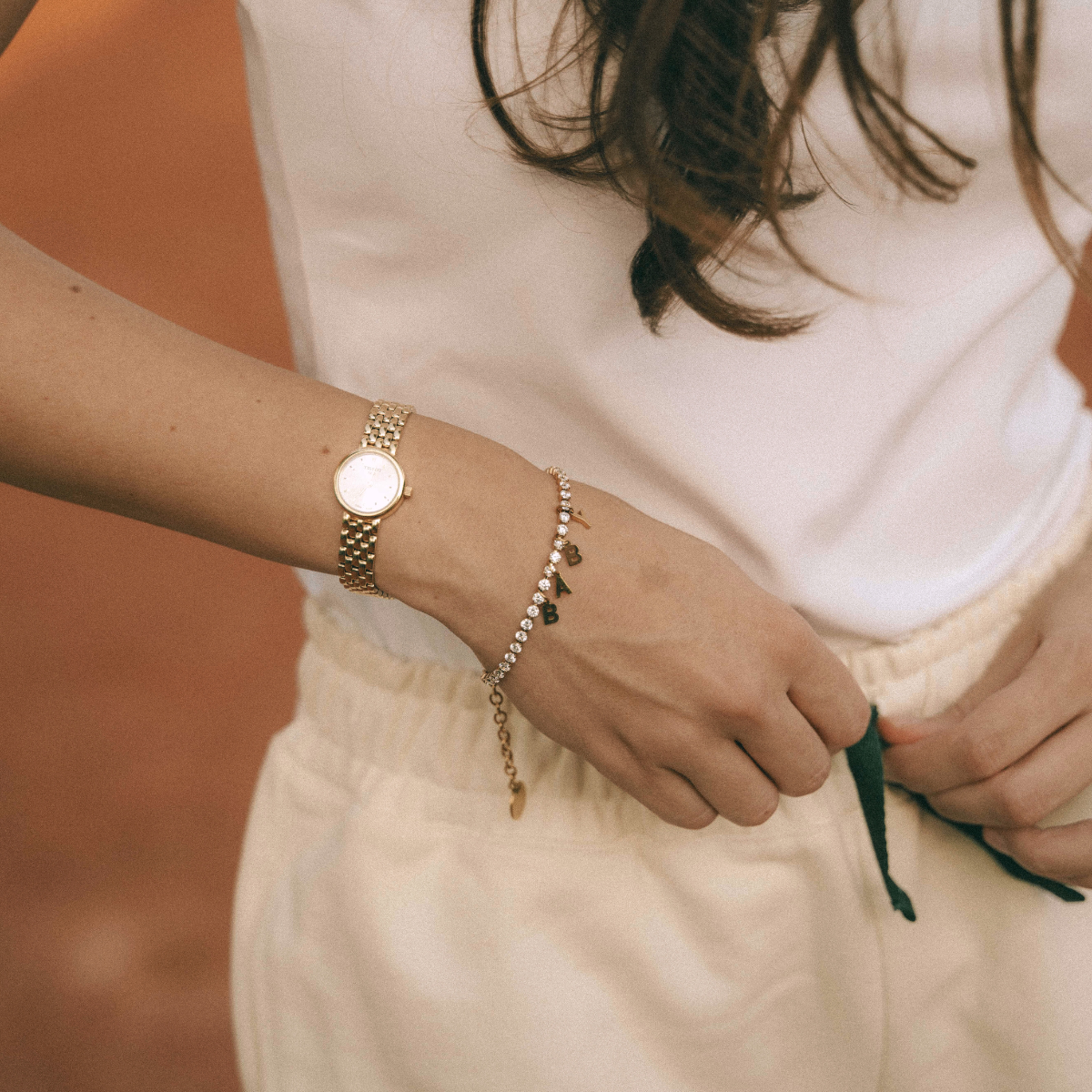 Woman wearing a gold watch and DIY tennis bracelet with zirconia stones and personalized letter charms, styled with casual beige pants