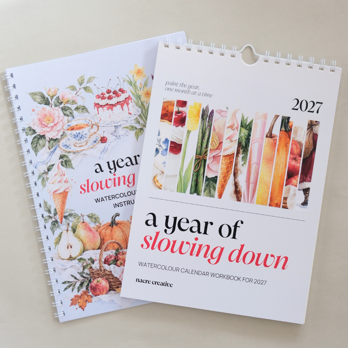 Two spiral-bound watercolour calendar workbooks titled "a year of slowing down" for 2027, shown on a light beige background. One cover features floral and fruit illustrations, and the other features vertical strips of colorful food and drink imagery.