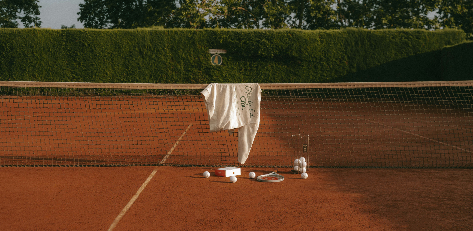 Tennis racket and balls on a clay tennis court with a net in the foreground featuring a tennis bracelet kit
