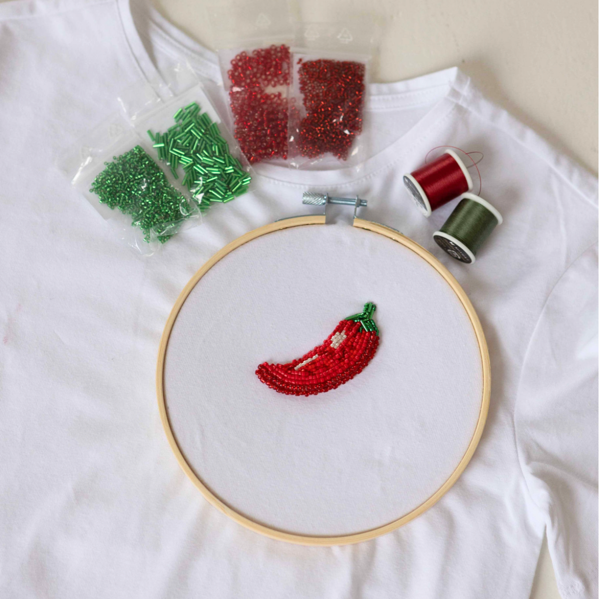 Embroidery kit including a white fabric with a chili pepper design, red and green Miyuki beads, embroidery thread, a needle, and an embroidery hoop.