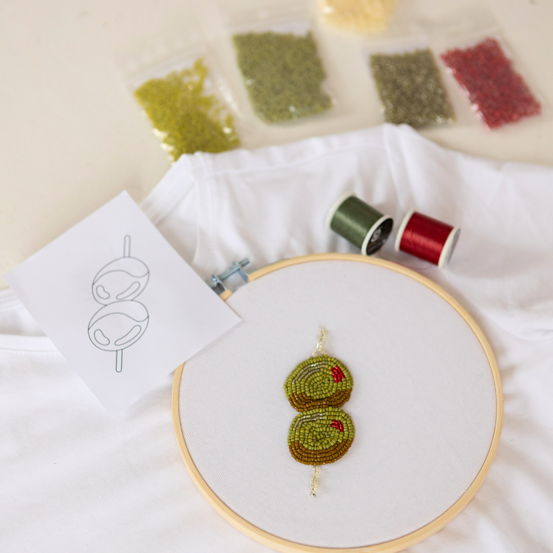 Embroidery kit contents displayed including a piece of fabric with beaded embroidery in progress, a hoop, embroidery thread, and a pattern template. DIY olives beaded embroidery kit with Miyuki seed beads, hoop, and pattern template.