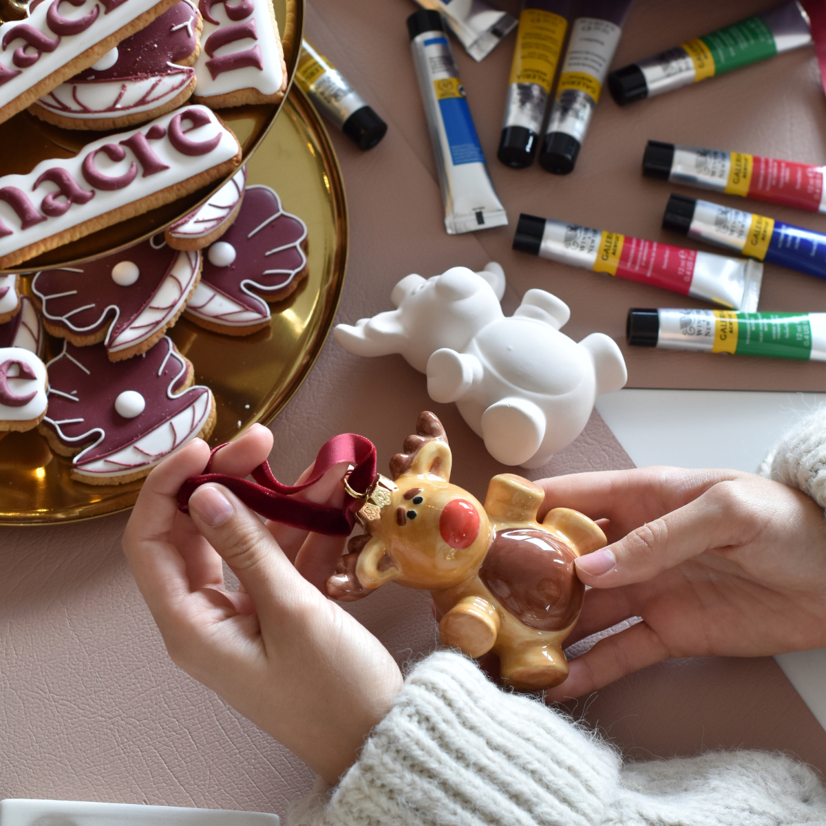Person decorating a small reindeer figurine with paint tubes in the background
