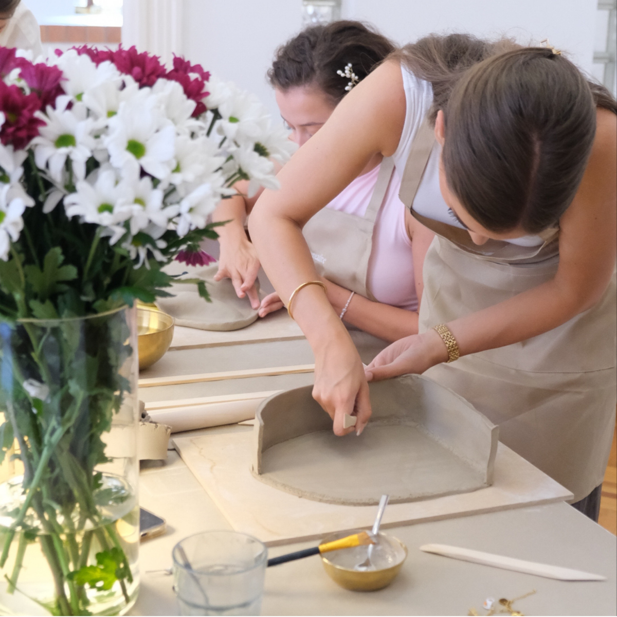 Two women working with clay on a pottery wheel in a bright studio.