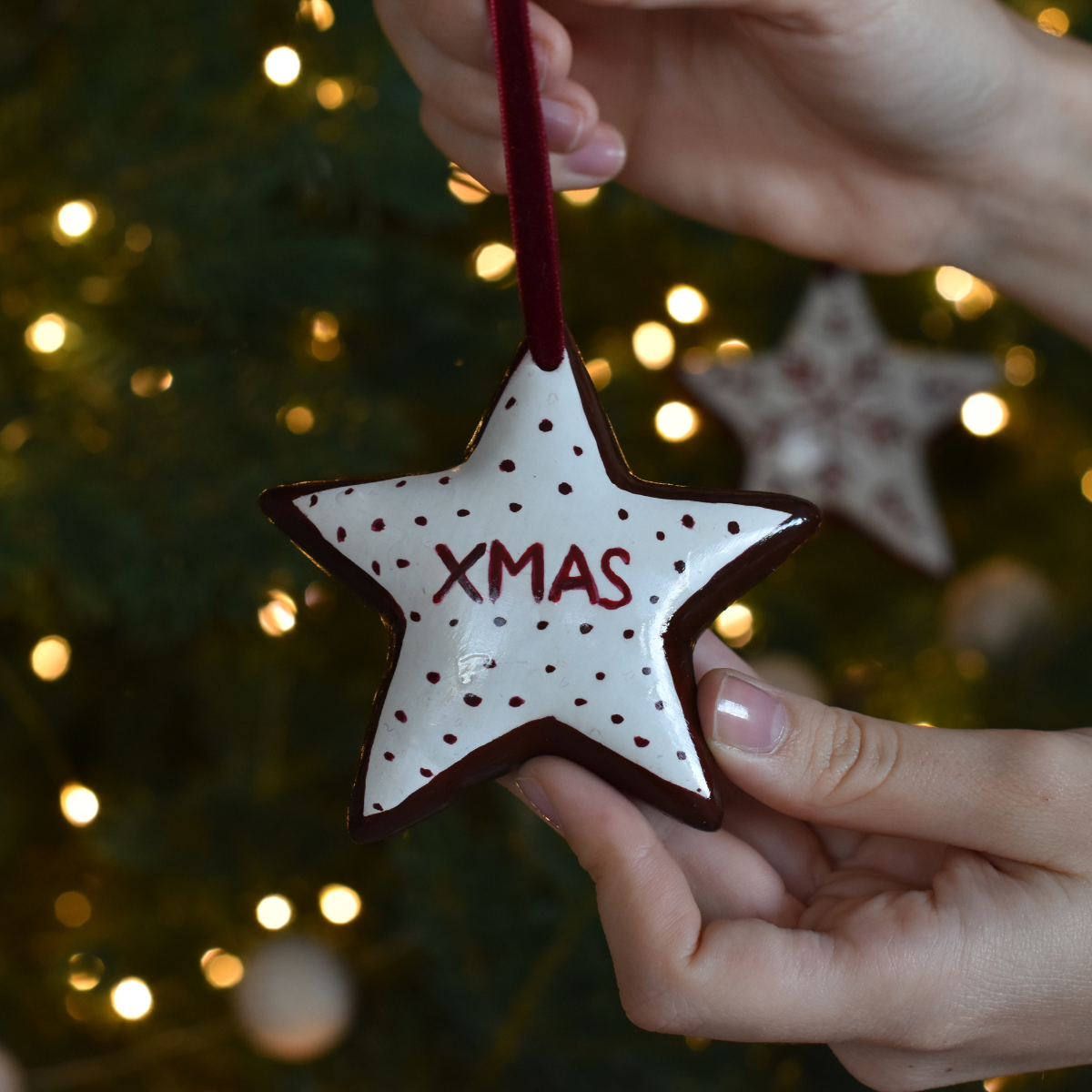 Star-shaped Christmas ornament with 'XMAS' text held by a hand against a blurred Christmas tree background.